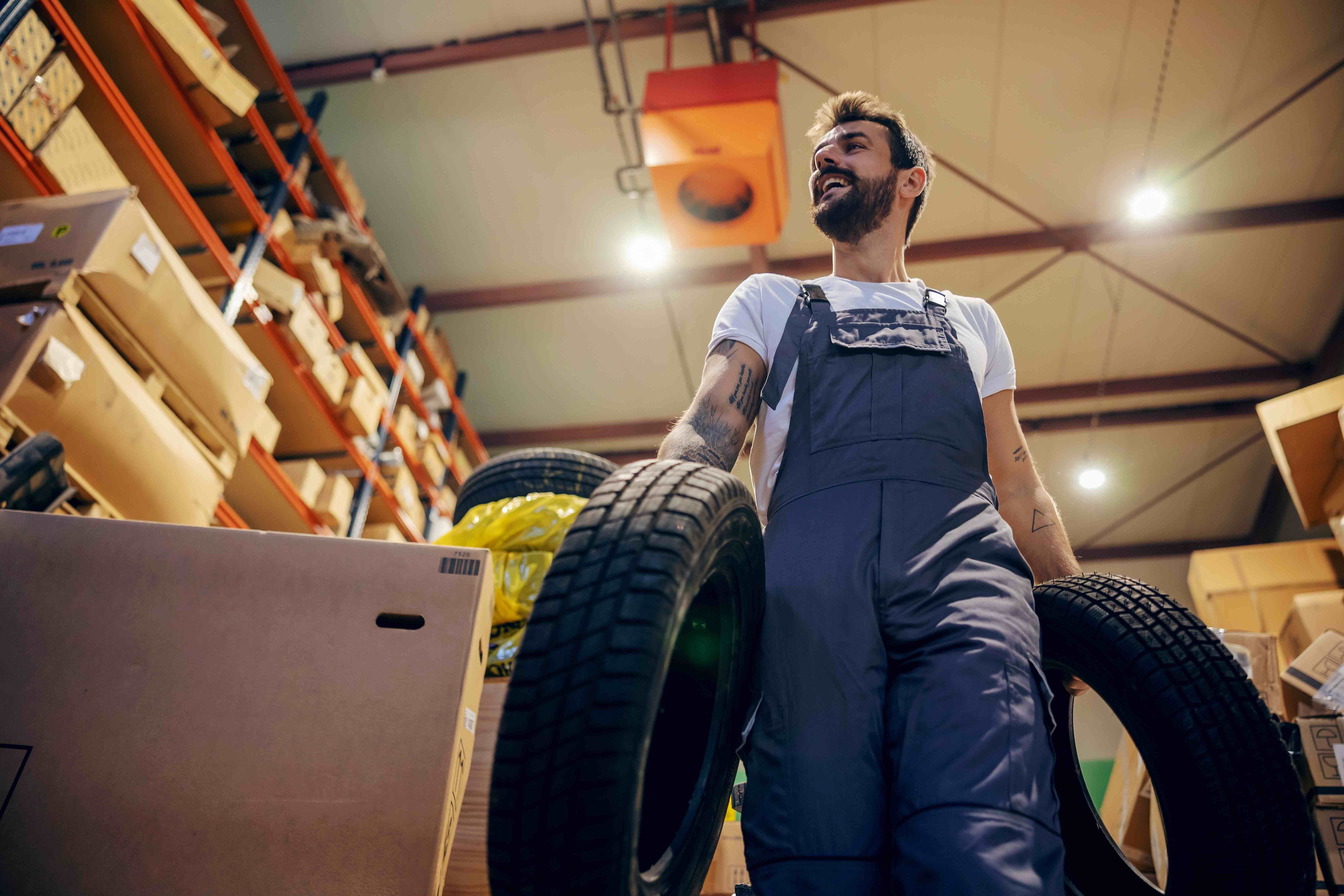 Low angle view of tattooed bearded hardworking smiling employee relocating tires while walking in storage of import and export firm.